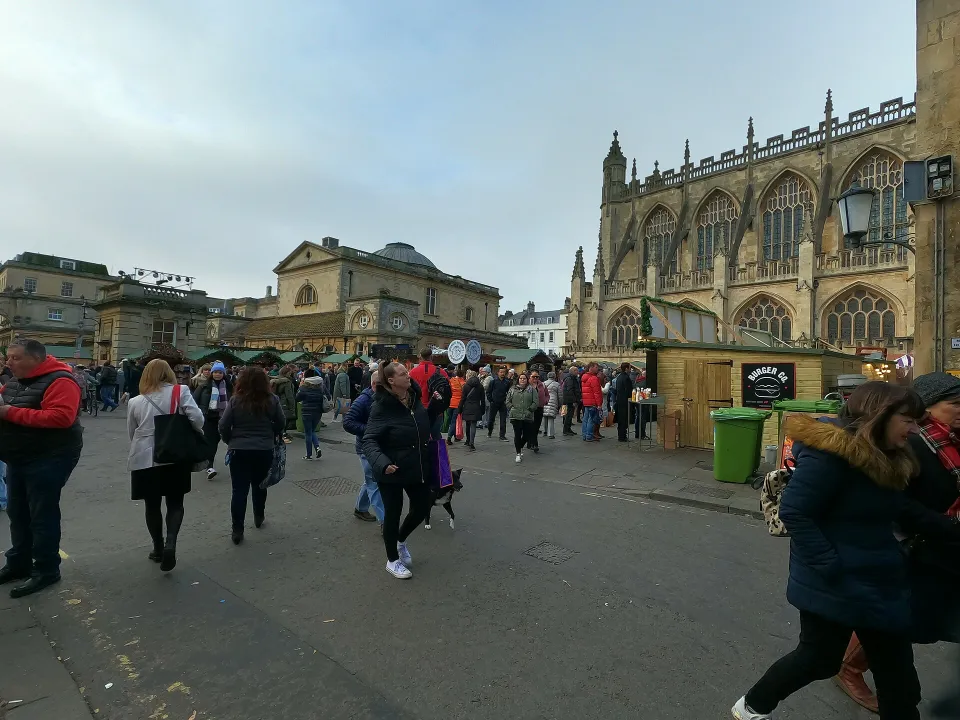 A crowd walks between green-roofed wooden chalets on a paved square with the side of Bath Abbey rising behind.