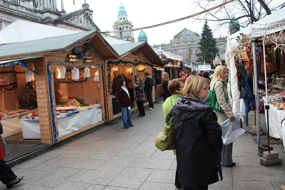 Shoppers browse a row of wooden chalets on a paved path, with the green copper domes of Belfast City Hall behind.
