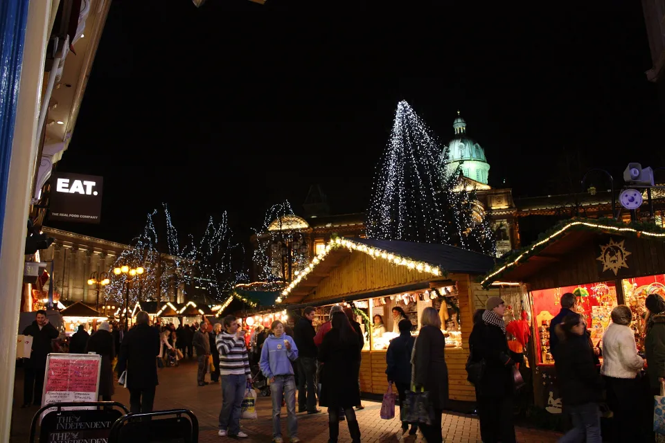 Wooden stalls edged with lights and a crowd of shoppers at night, with illuminated trees and a lit dome in the background.