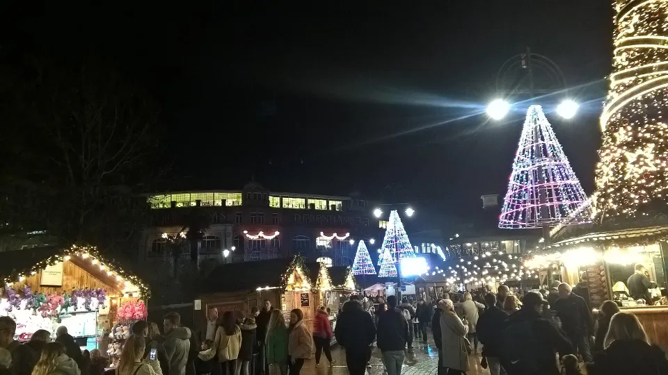 A crowd between wooden chalets at night, with cone-shaped trees of pink and white lights rising behind.