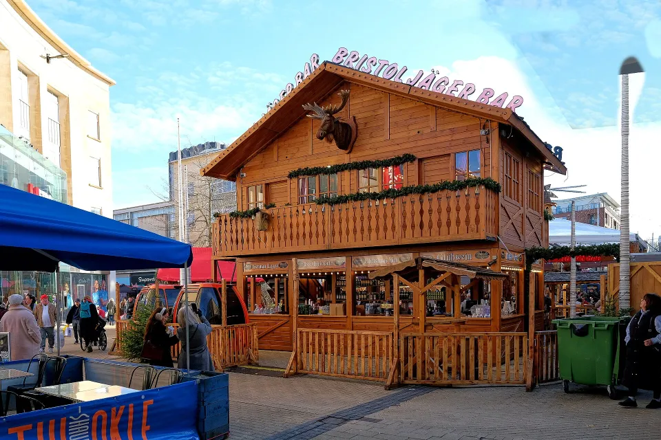A two-storey wooden alpine chalet bar with a balcony and a moose head, signed Bristol Jäger Bar, in a pedestrian shopping street under a blue sky.