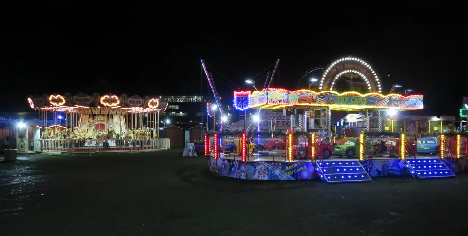 Brightly lit fairground rides, including a carousel and a children's car ride, on dark grass at night.