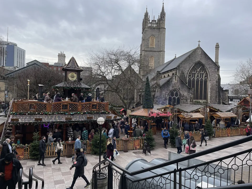 Seen from above, a two-level wooden bar and rows of chalets fill a paved street busy with shoppers, with the tower of St John's Church behind.