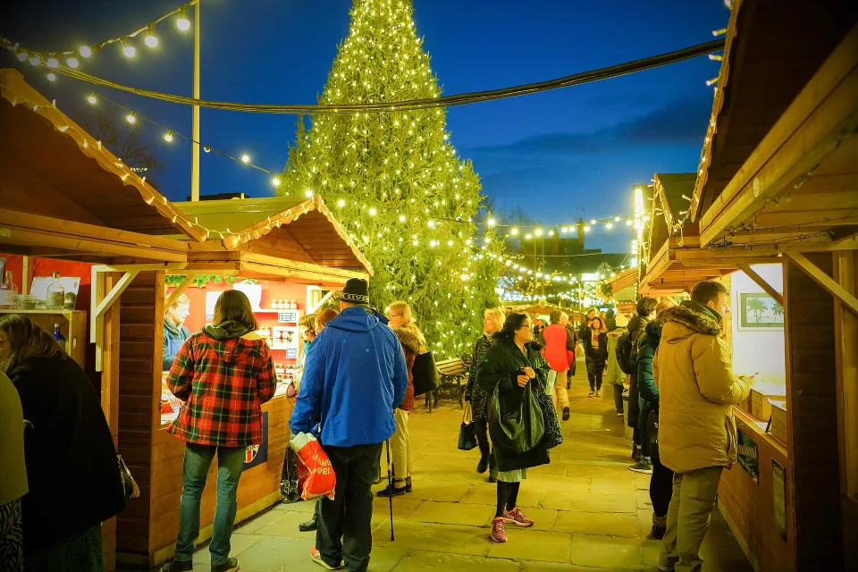 Shoppers walk between wooden chalets strung with festoon lights at dusk, with a tall Christmas tree covered in white lights behind.
