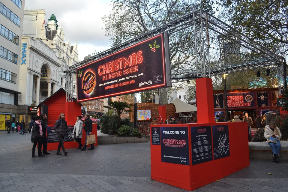 A gantry sign reading Christmas in Leicester Square over red entrance kiosks and a red hut, with people walking through the square.