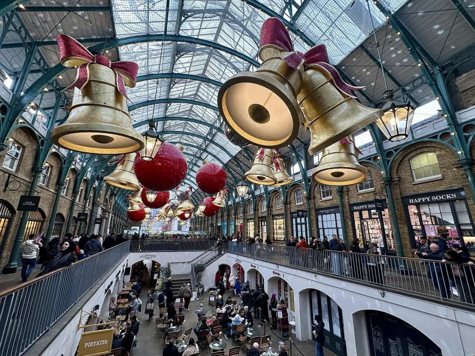 Giant golden bells with red bows and red baubles hang under the glass and iron roof of the market building, above a crowded lower courtyard.
