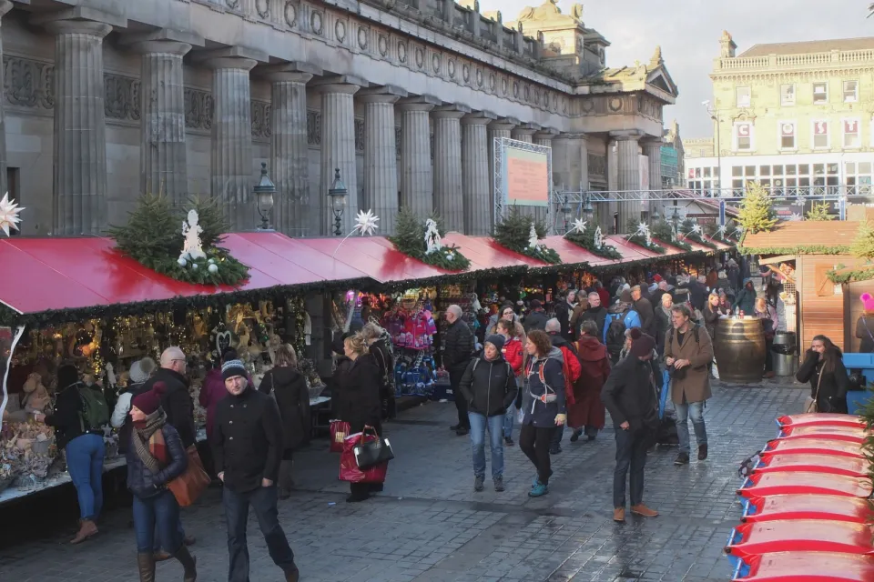 A row of red-roofed market stalls trimmed with greenery beside the columned Royal Scottish Academy, with shoppers walking past on wet paving.