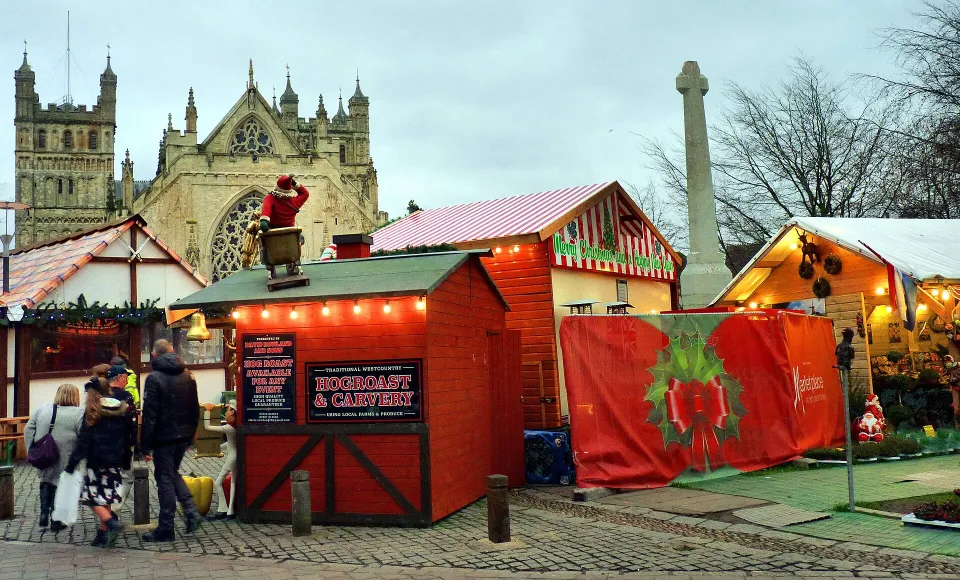 Red and striped wooden chalets, one topped with a Father Christmas figure, on a cobbled green with Exeter Cathedral behind.