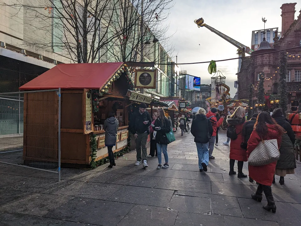 People walk past a red-roofed wooden chalet on a paved square, with more stalls, fairground rides and a red sandstone building beyond.