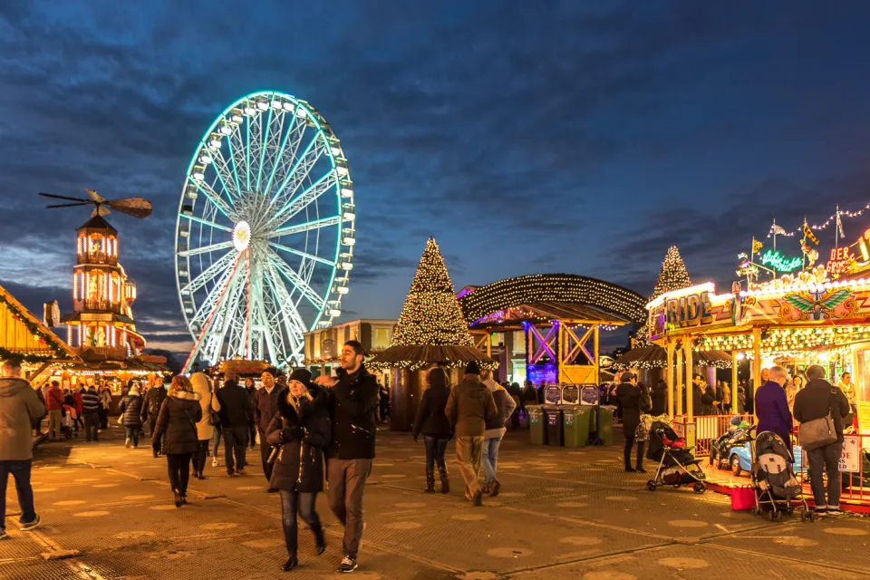 Visitors walk at dusk between lit stalls and rides, with a Christmas pyramid, a cone of lights and a large illuminated observation wheel behind.