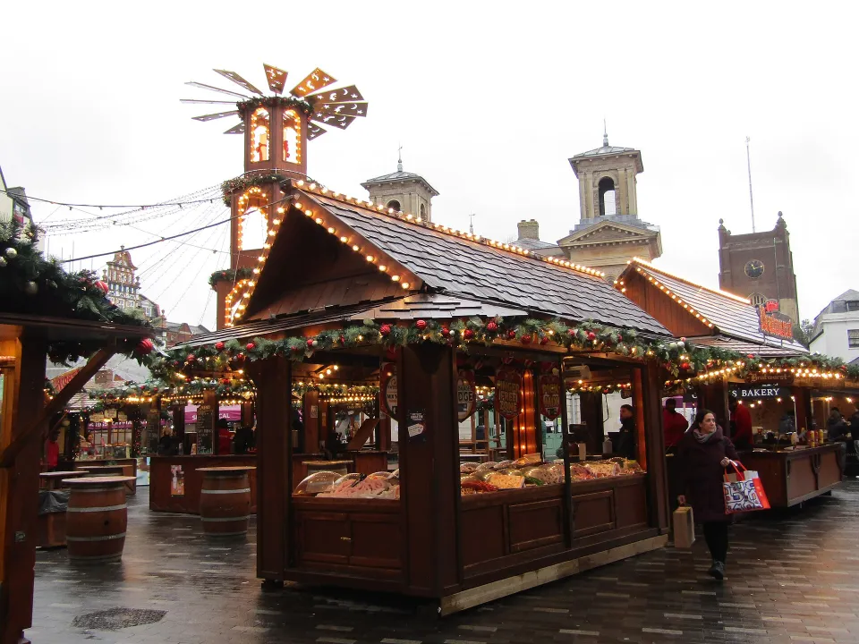 Dark wooden chalets with lit roof edges and garlands on wet paving, with a Christmas pyramid and the towers of the Market House behind.