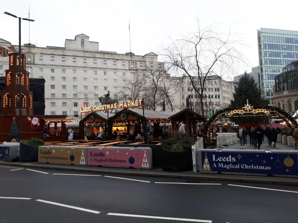 Wooden chalets, a lit entrance arch and a sign reading Leeds Christmas Market in a city square, seen across a road with large stone buildings behind.