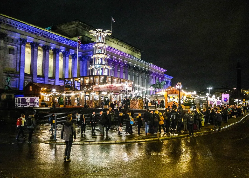 Market chalets and a tall Christmas pyramid stand lit up at night in front of the columns of St George's Hall, with people crossing the wet road.