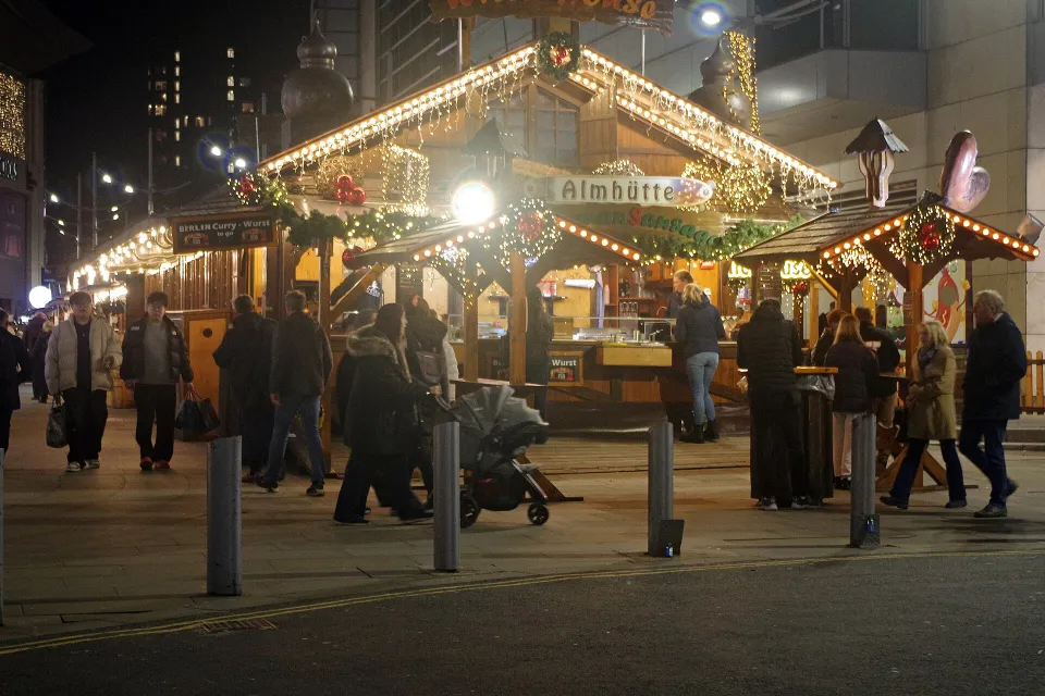 Wooden food and drink chalets outlined in white lights at night, with people walking past and standing at tables.