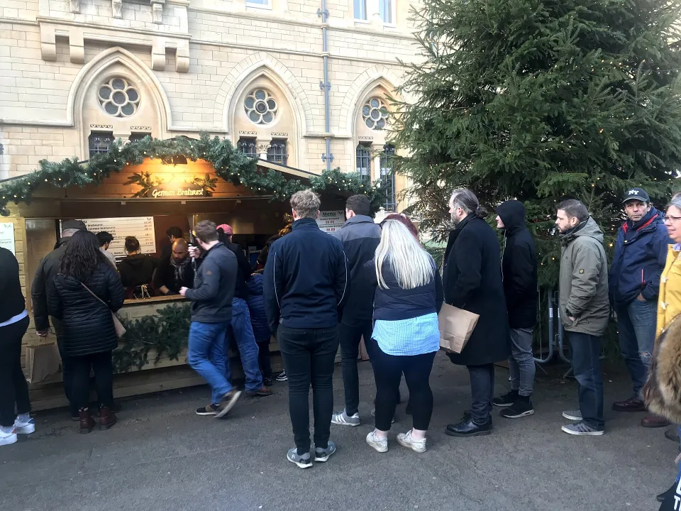A queue of people in coats, seen from behind, waits at a wooden food chalet trimmed with fir garlands beside a large Christmas tree.