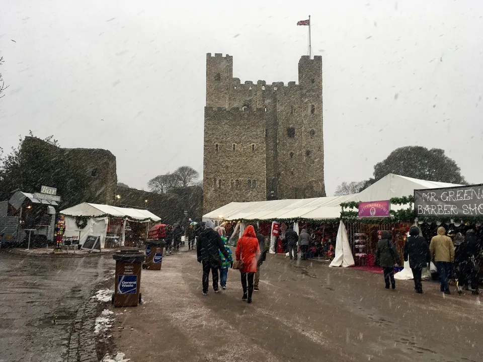 Visitors in winter coats walk past white market tents and stalls in falling snow, with the stone keep of Rochester Castle behind.