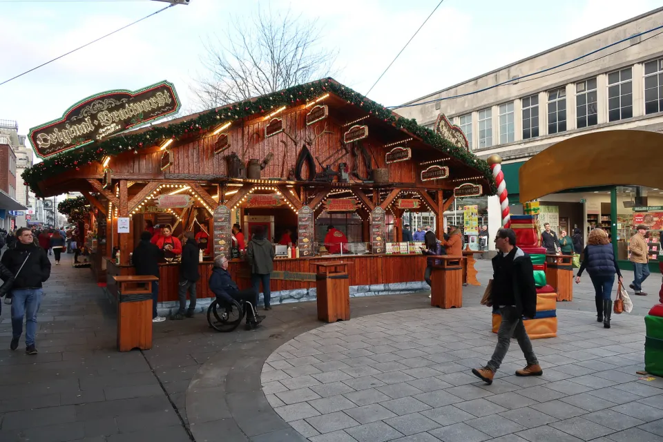 A large open-sided wooden food chalet decorated with garlands and lights in a pedestrian shopping street, with customers at the counter.