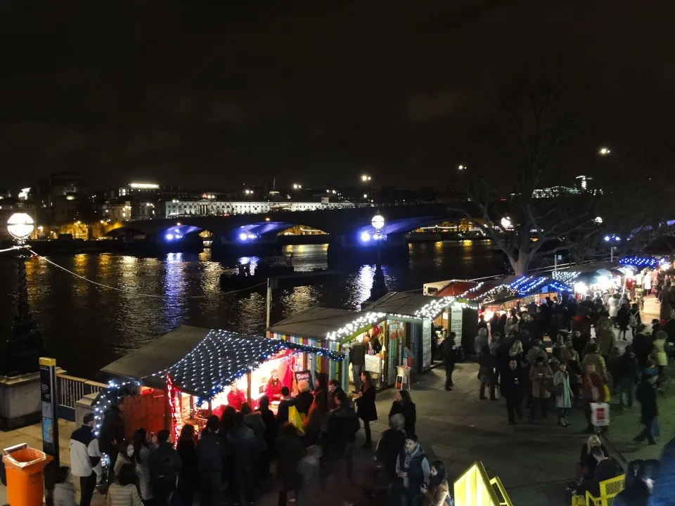 Seen from above at night, a line of chalets with fairy-lit roofs runs along the Thames path, crowded with visitors, with a lit bridge across the river.