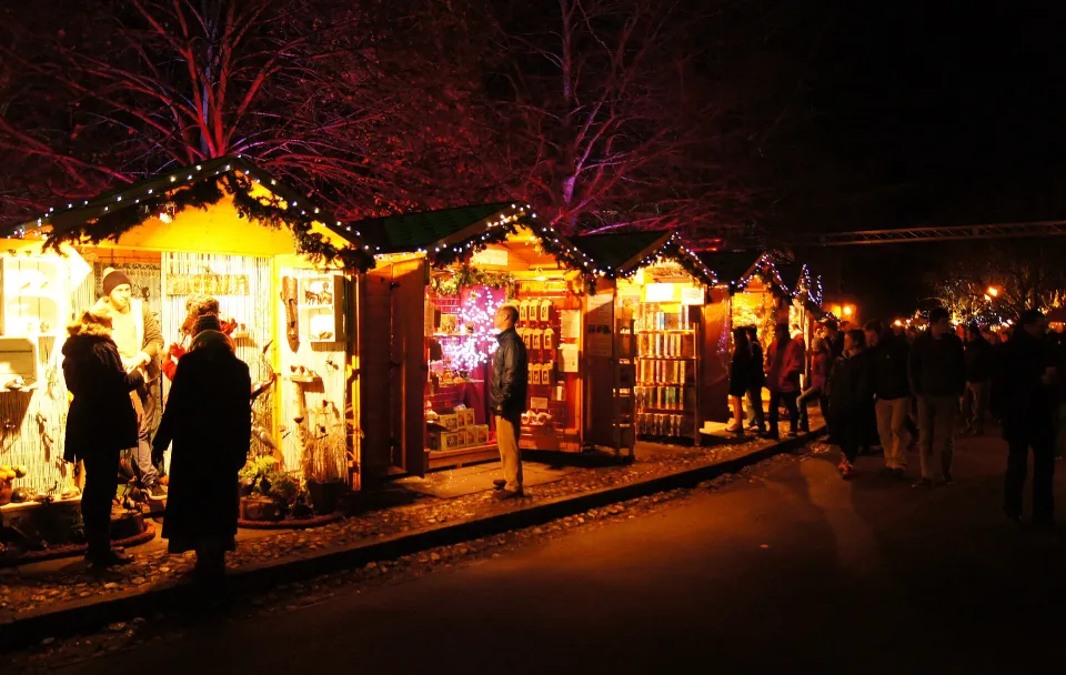 A row of small wooden chalets glowing with warm light after dark, with visitors browsing and trees lit red behind.