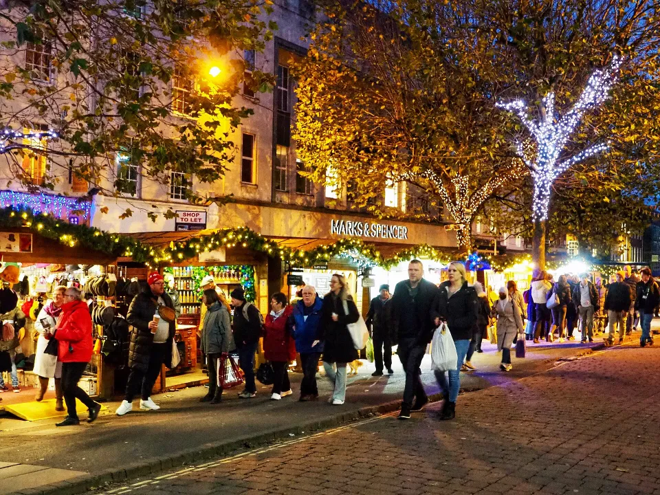 Shoppers walk past wooden stalls hung with garlands and lights at dusk, under trees wrapped in white fairy lights.